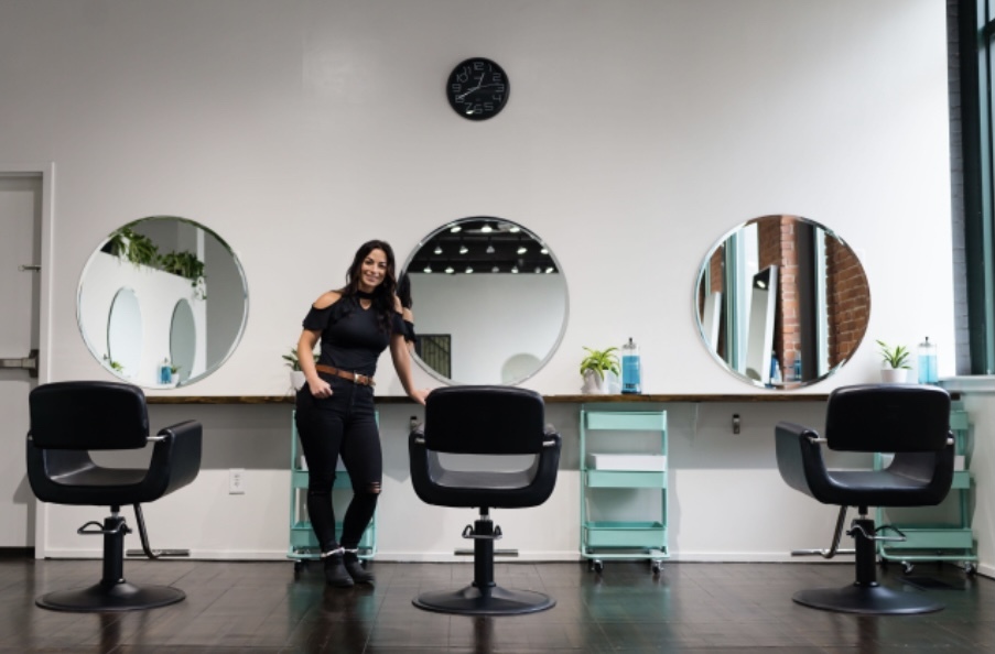 A stylist stands smiling in a modern hair salon between three black salon chairs positioned in front of round mirrors